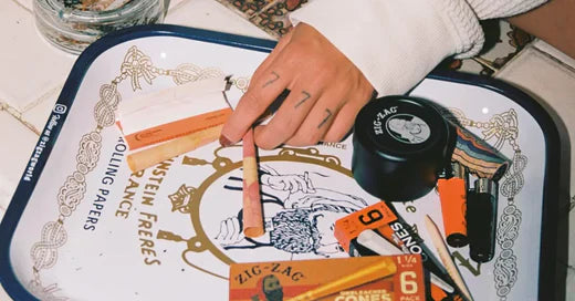 Smoking tools arranged on a decorative tray, including rolling papers, pre-rolled cones, a grinder, lighters, and a hand holding a rolled paper.