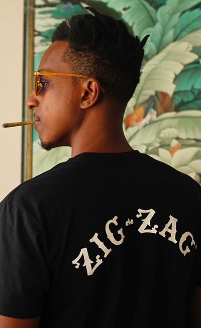 Person wearing a black Zig-Zag t-shirt with bold white lettering, holding a rolled product against a tropical backdrop.