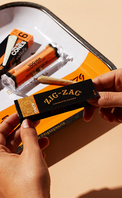 Hands holding a pack of Zig-Zag King Size cigarette papers, with cones and rolling accessories on a tray in the background.