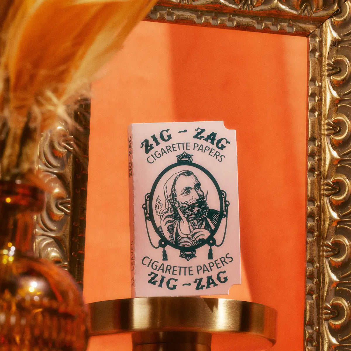 Zig-Zag cigarette papers displayed on a golden shelf with a decorative frame and warm orange background.