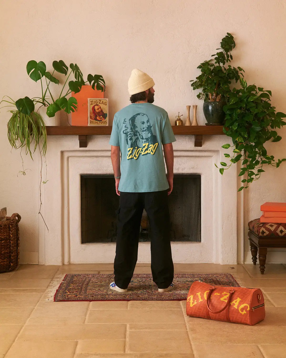 Man wearing a teal Zig-Zag t-shirt with a large back graphic, black pants, and a cream beanie, standing in a cozy room.