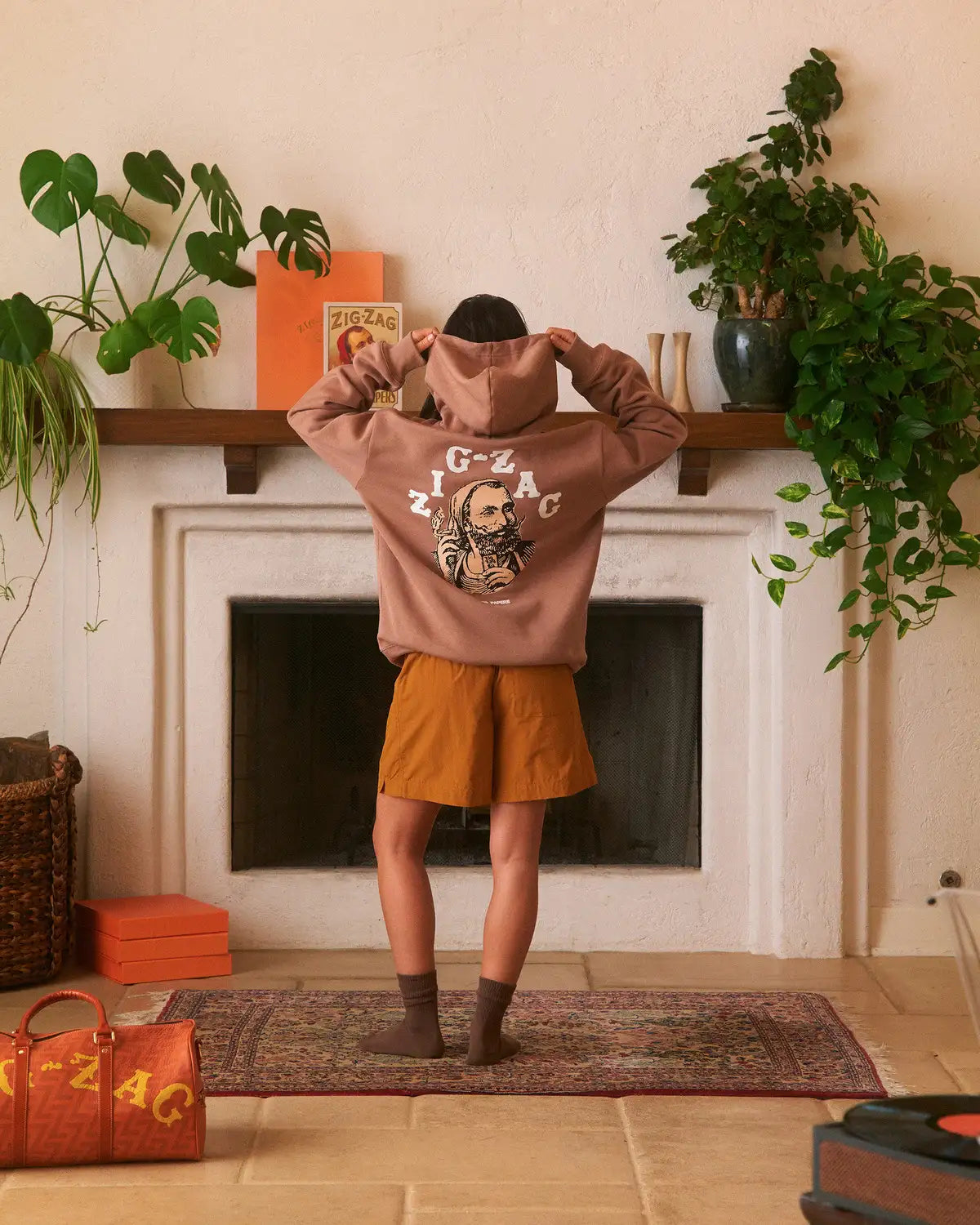 Woman showing the back graphic of a brown Zig-Zag hoodie, paired with orange shorts, in a cozy room with plants and decor.