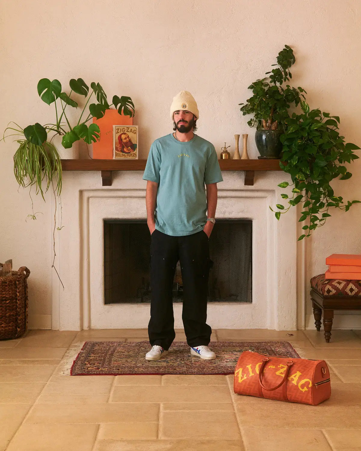 Man wearing a teal Zig-Zag t-shirt, black pants, and a cream beanie, standing in a cozy room with plants and decor.