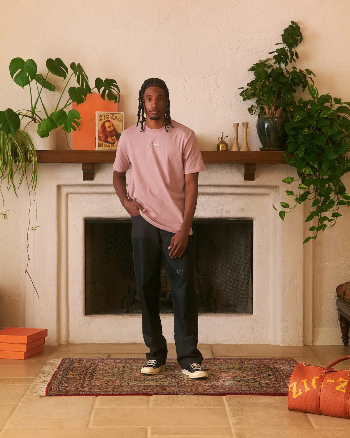 Man wearing a mauve Zig-Zag t-shirt and black pants, standing in a cozy room with plants and decor.