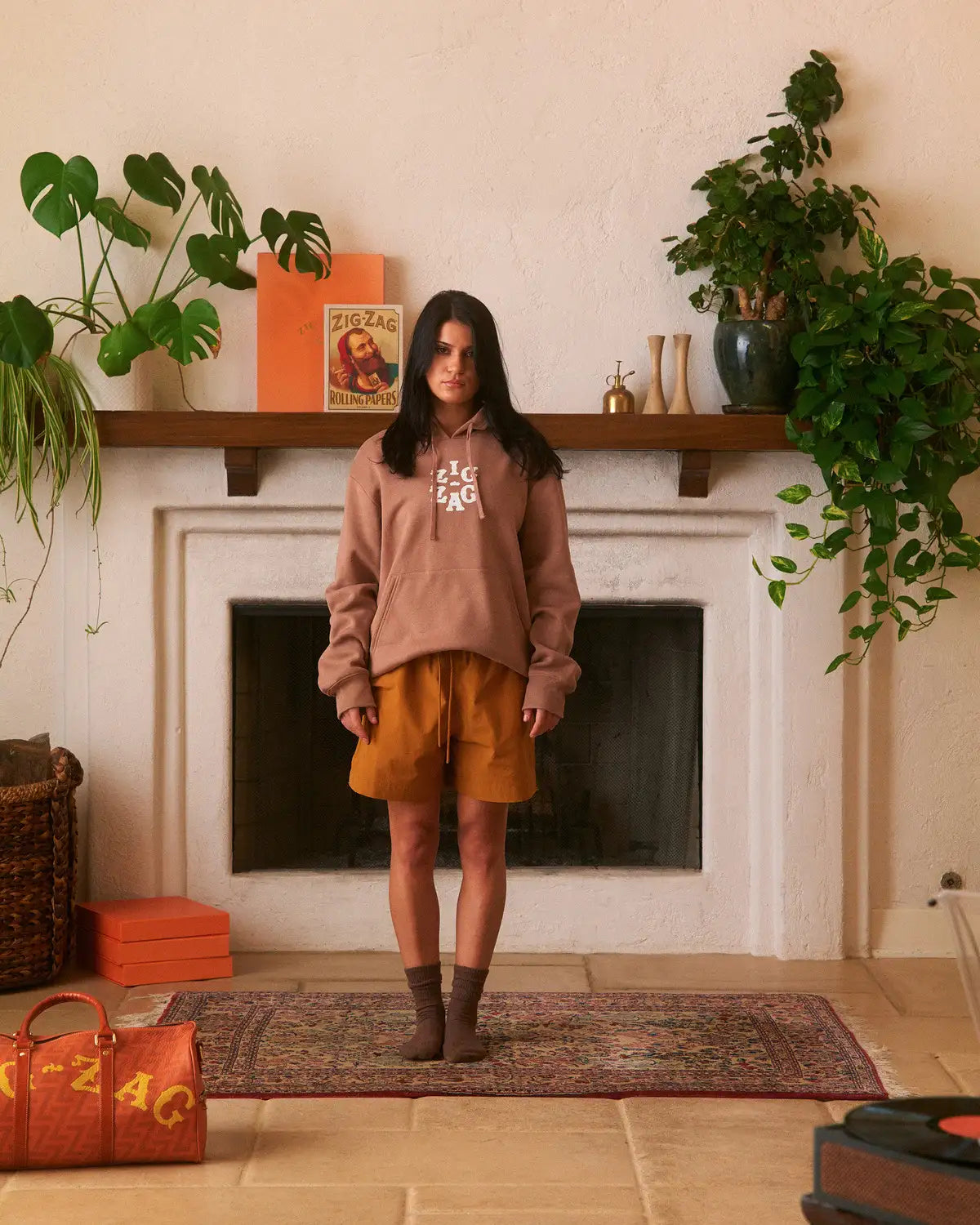 Woman wearing a brown Zig-Zag hoodie and orange shorts, standing in a cozy room with plants and decor.